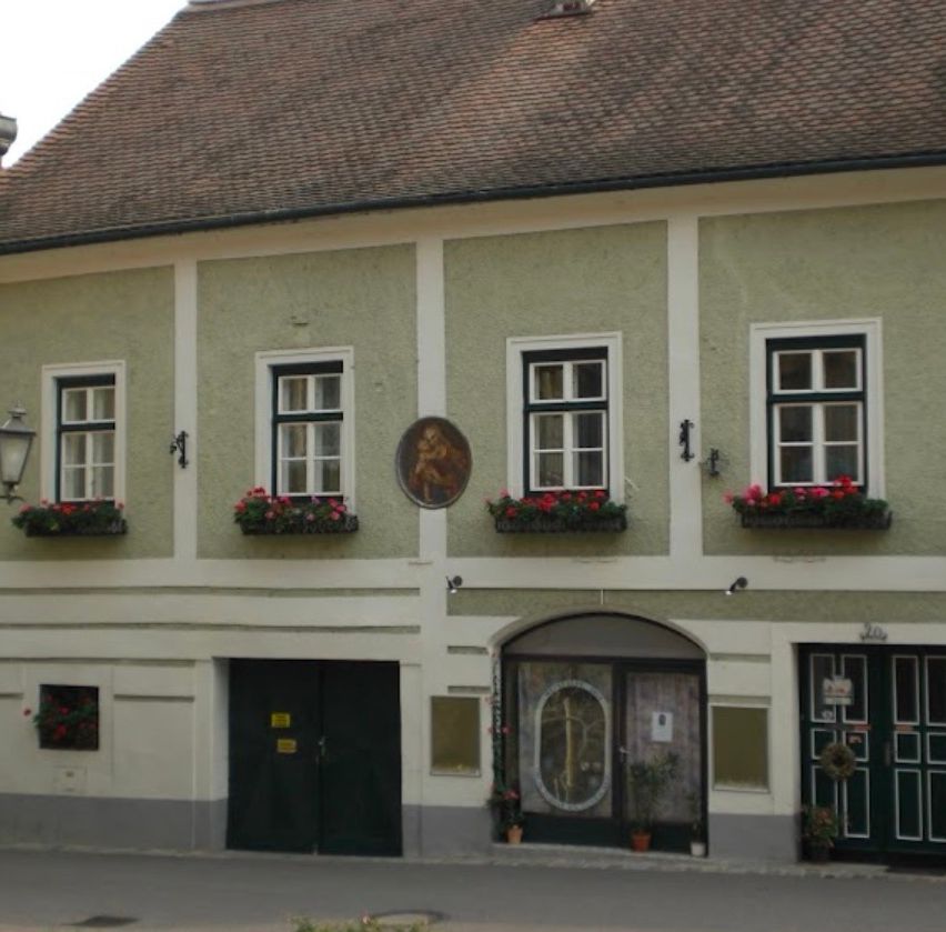 Ein traditionelles europäisches Gebäude mit einem braunen Dach und grünen Wänden. Es gibt drei Fenster mit Blumenkästen darunter und eine zentrale Tür. Zwei kleinere Türen befinden sich auf beiden Seiten. Eine Plakette ist über der zentralen Tür angebracht.