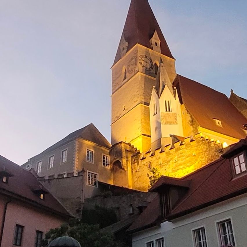Eine historische Kirche mit Kirchturm und Uhr wird gegen einen klaren blauen Himmel beleuchtet. Die Kirche ist von Gebäuden mit braunen Dächern und Fenstern umgeben.