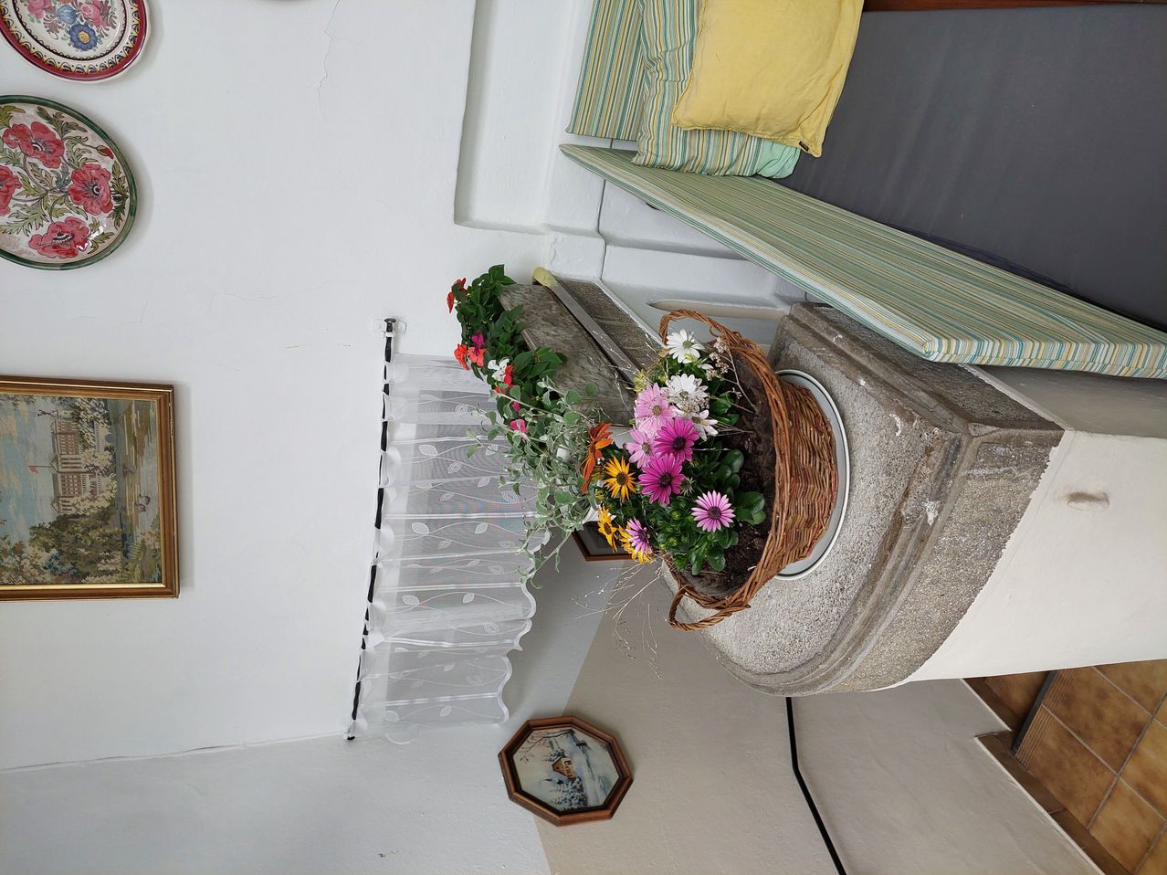 Auf einem Steinpodest steht ein Korb aus Weidenflechtwerk, gefüllt mit leuchtenden rosa, gelben und weißen Blumen. Dahinter schmückt eine gerahmte Bild und ein weißer Vorhang die Wand.