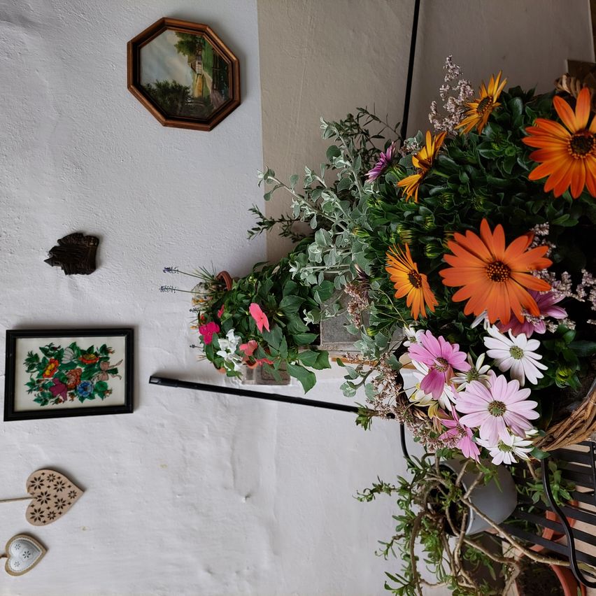 An einer weißen Wand hängt ein großer Blumenstrauß. Ein achteckiger Rahmen und ein kleines gerahmtes Bild schmücken die Wand. Ein holzherz und ein schwarzer Gegenstand hängen unter dem Rahmen.