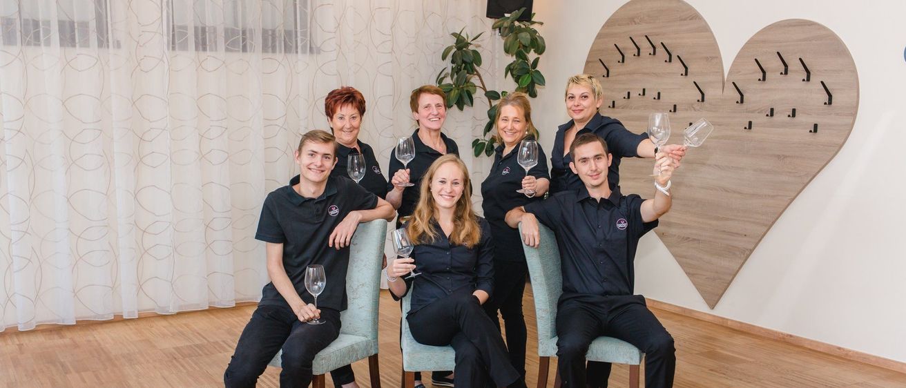 Eine Gruppe von sieben Erwachsenen, sowohl Männer als auch Frauen, gekleidet in übereinstimmenden schwarzen Uniformen, sitzen auf Stühlen in einem Raum und posieren für ein Foto. Sie halten alle Weingläser.