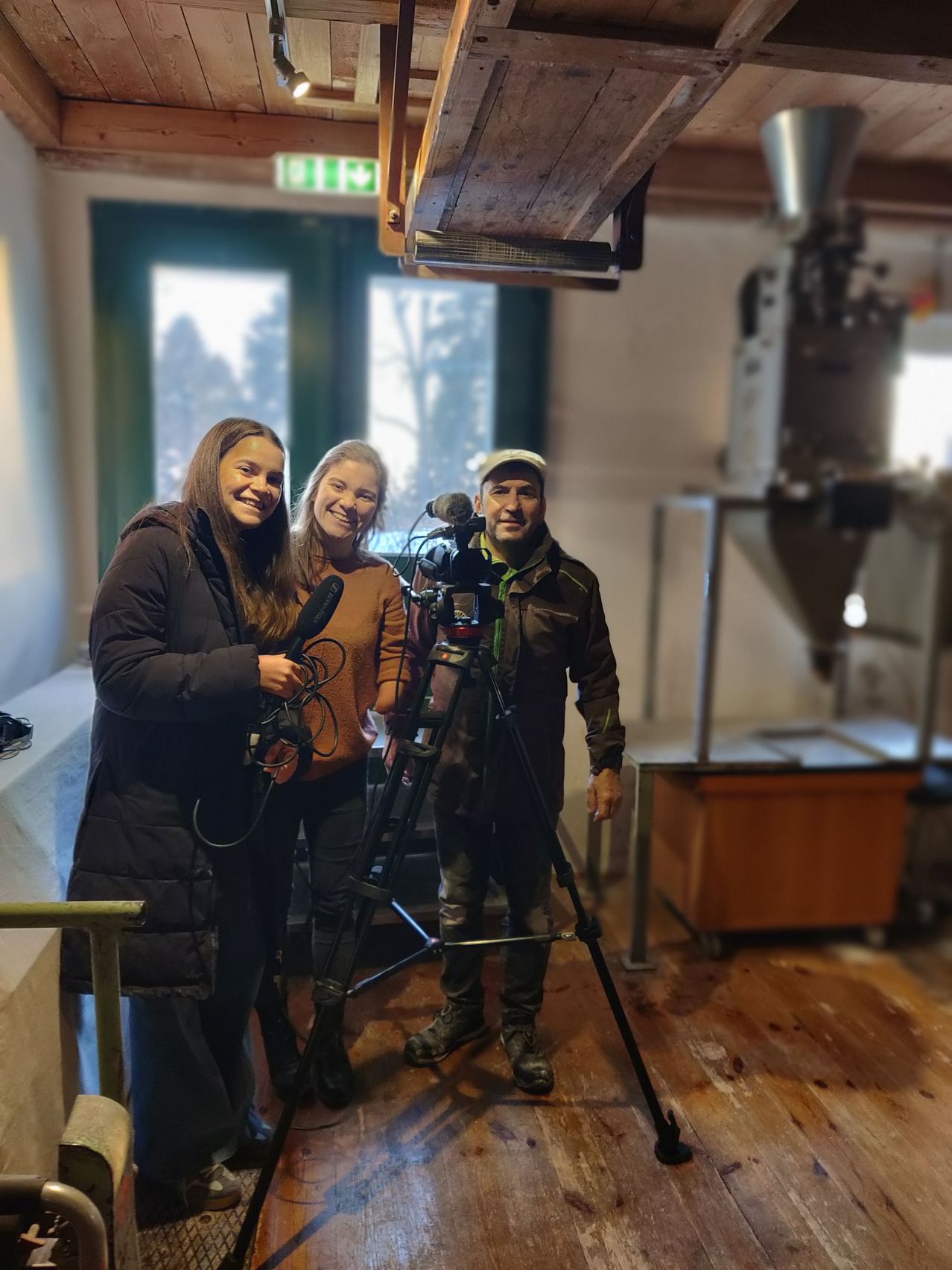 Three people are standing indoors, smiling, with a camera and microphone on a tripod between them. The room has wooden floors and a machine with a large pipe in the background.