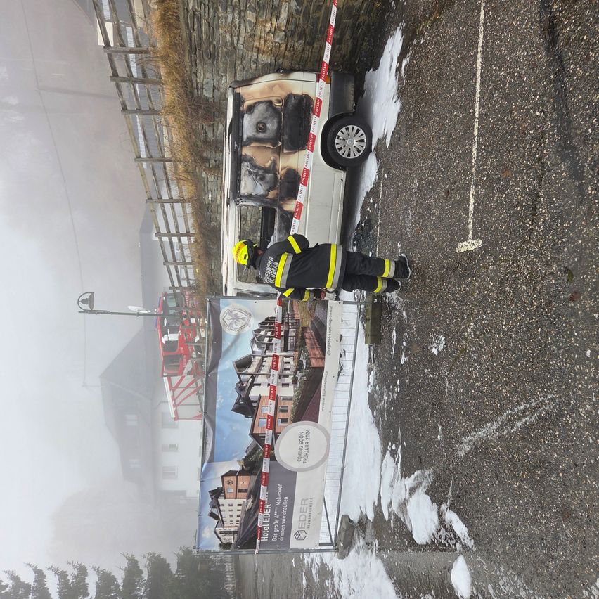 Ein Feuerwehrmann in Schutzkleidung steht neben einem ausgebrannten Van auf einer verschneiten Straße. In der Nähe ist ein Poster zu sehen, das EDELER bewirbt.