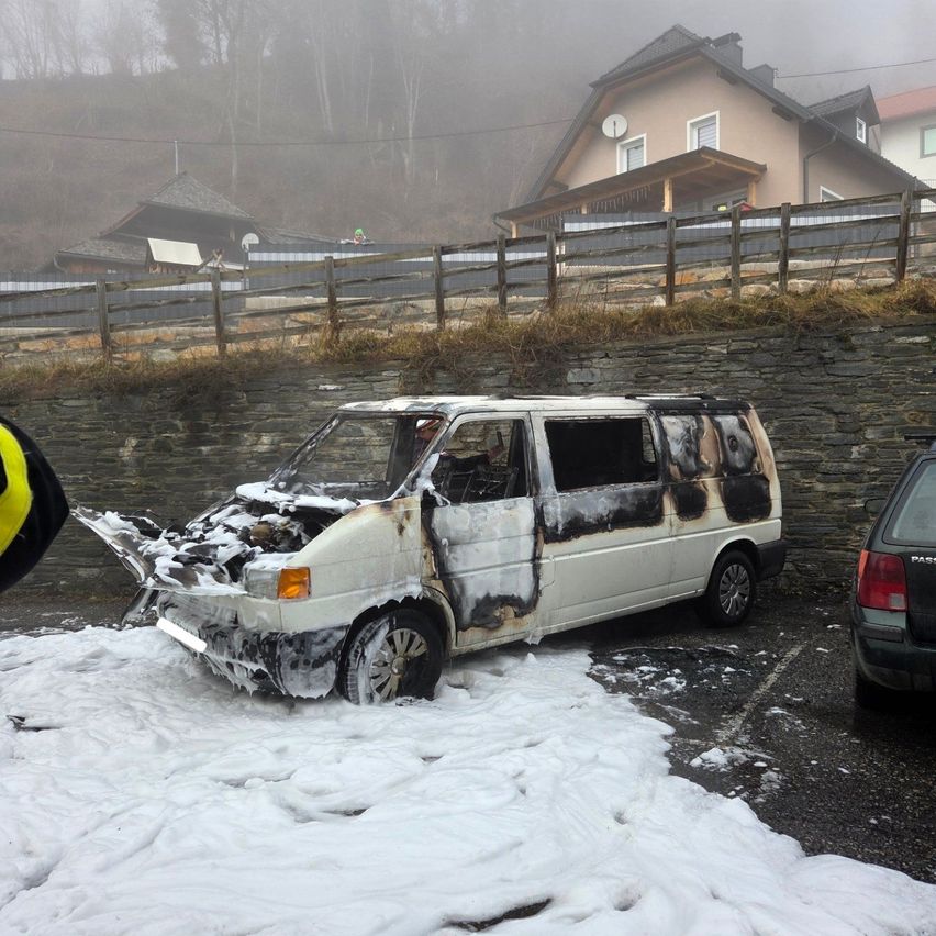 Ein ausgebrannter weißer Van steht auf einem Parkplatz. Er hat eine kaputte Front und ein beschädigtes Dach. Schnee bedeckt den Boden. Ein Haus und ein Zaun sind im Hintergrund.