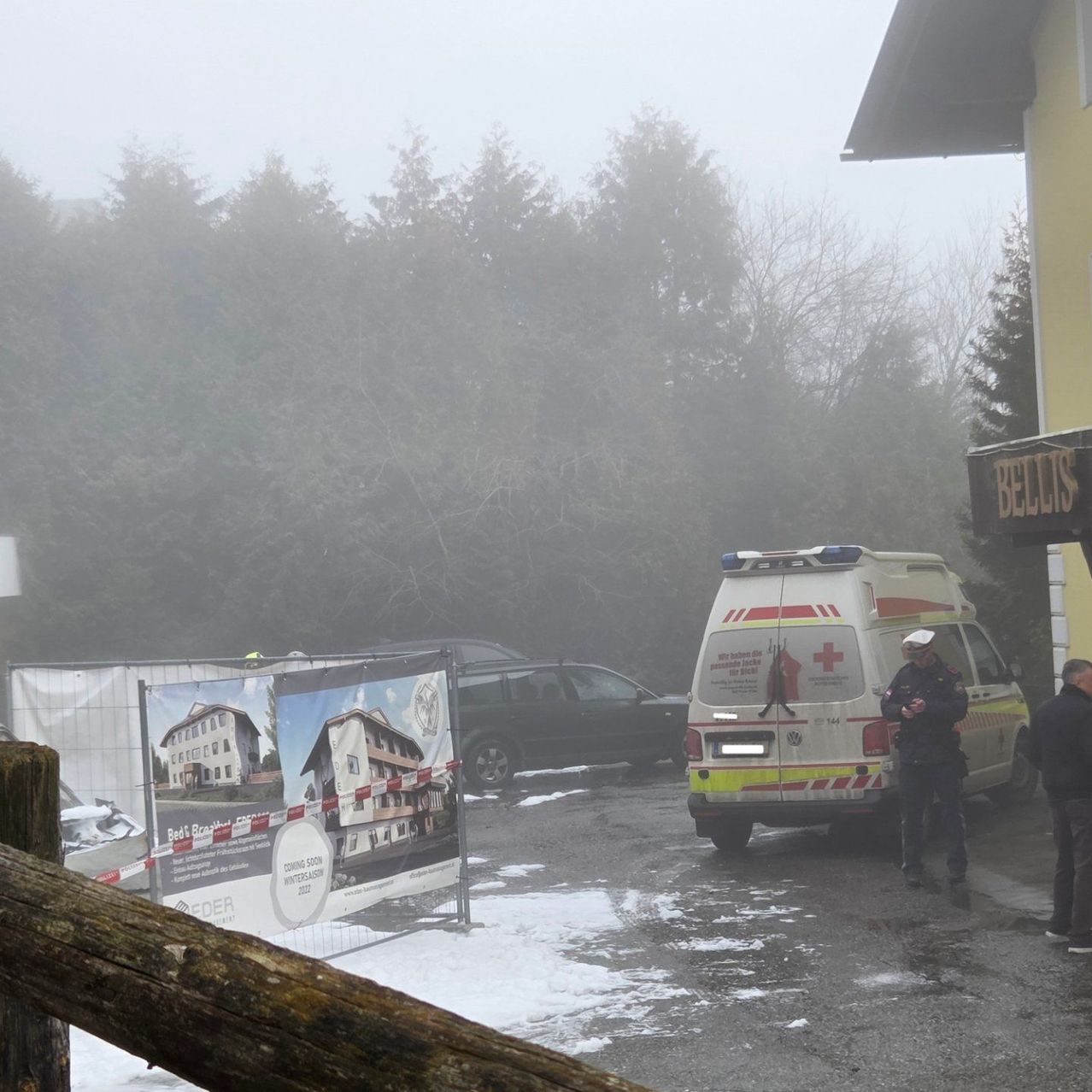 Ein Krankenwagen ist am Straßenrand geparkt. Zwei Personen stehen neben dem Krankenwagen. Vor dem Krankenwagen befindet sich ein Zaun mit einem Schild. Ein Gebäude ist in der Ferne sichtbar.