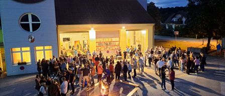 Eine große Gruppe von Menschen versammelt sich bei Nacht vor einem Gebäude mit Turm. Viele stehen und unterhalten sich, einige um ein Feuer herum. Es gibt aufgestellte Bänke und Tische.
