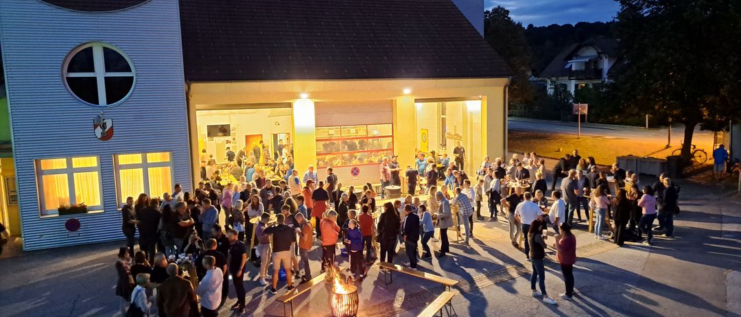 Eine große Gruppe von Menschen versammelt sich bei Nacht vor einem Gebäude mit Turm. Viele stehen und unterhalten sich, einige um ein Feuer herum. Es gibt aufgestellte Bänke und Tische.