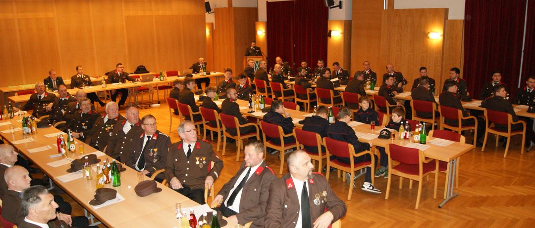 Eine Gruppe von Menschen in Uniform sitzt um Tische in einem Raum, einige trinken und andere schauen sich Papiere an. Ein Mann vorne trägt einen Hut und eine Medaille.