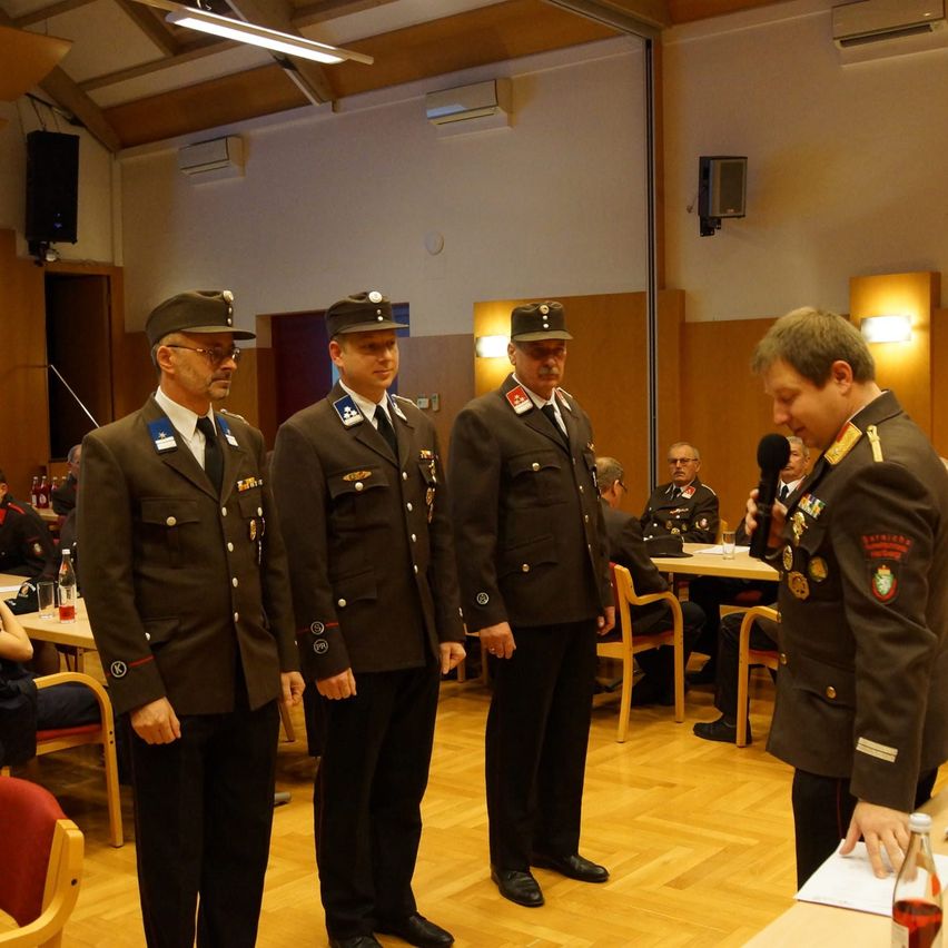 Vier Männer in Militäruniformen stehen in einem Raum mit Holzboden. Sie nehmen anscheinend an einer formellen Veranstaltung teil. Ein Mann mit einem Mikrofon steht vor ihnen.