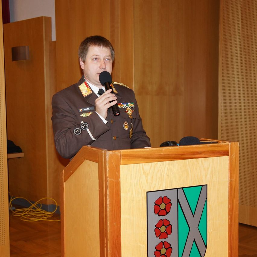 Ein Mann in Uniform spricht in ein Mikrofon an einem Podium. Das Podium zeigt ein Logo mit roten Blumen und einem Buchstaben 'X'. Dahinter befindet sich eine Holzwand.