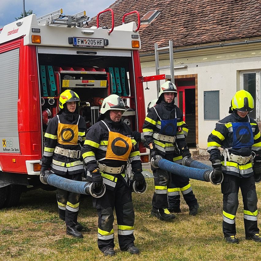 Five firefighters in uniform with helmets stand in front of a fire truck with an open door. They hold hoses. The building behind has a red door and windows.