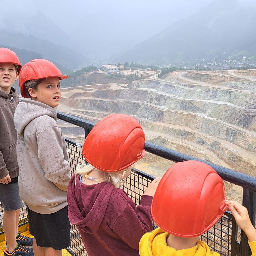 Vier Kinder mit roten Helmen stehen auf einer Plattform und blicken auf eine große offene Grube. Die Mine ist von Bergen und Nebel umgeben.