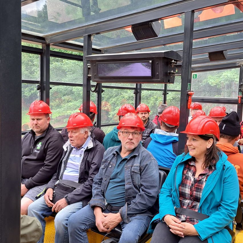 Eine Gruppe von Menschen sitzt in einem Bus und trägt rote Helme. Sie schauen nach vorne, möglicherweise auf einer Tour. Der Bus hat ein Glasdach und Fenster.