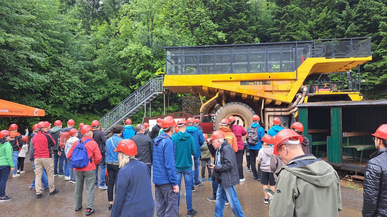 Eine Gruppe von Menschen mit roten Schutzhelmen steht in der Nähe eines großen gelben Muldenkipper-LKWs mit der Nummer HD 785. Der LKW ist auf einem Betonweg geparkt. Hinter dem LKW befindet sich eine Treppe mit Geländer. Im Hintergrund sind Bäume zu sehen.
