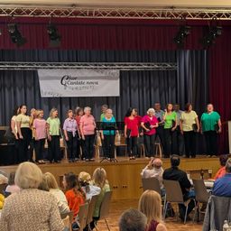 Ein Chor, 'Chor Cantate nova', tritt auf der Bühne auf. Zuhörer sitzen in Stühlen darunter, einige mit Getränken. Ein Banner mit Musiknoten hängt über der Bühne.