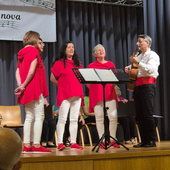 Vier Frauen in roten Outfits singen mit einem Mann, der eine Gitarre spielt, auf der Bühne. Ein Notenständer mit Noten steht vor ihnen. Ein Banner mit Musiknoten hängt im Hintergrund.