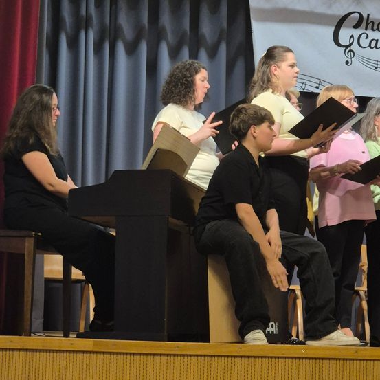 Eine Gruppe von Menschen steht auf der Bühne und führt ein Musikstück auf. Sie halten Notenblätter in den Händen und singen. Die Bühne ist gut beleuchtet und hat einen Hintergrund mit Noten.