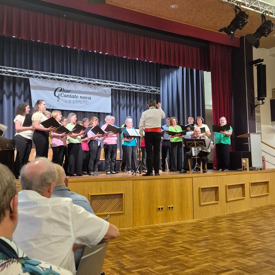 Ein Chor singt auf der Bühne mit Musikinstrumenten und einem Banner mit der Aufschrift 'Chor Cantante Nova'. Ein Publikum sitzt auf Stühlen vor der Bühne.