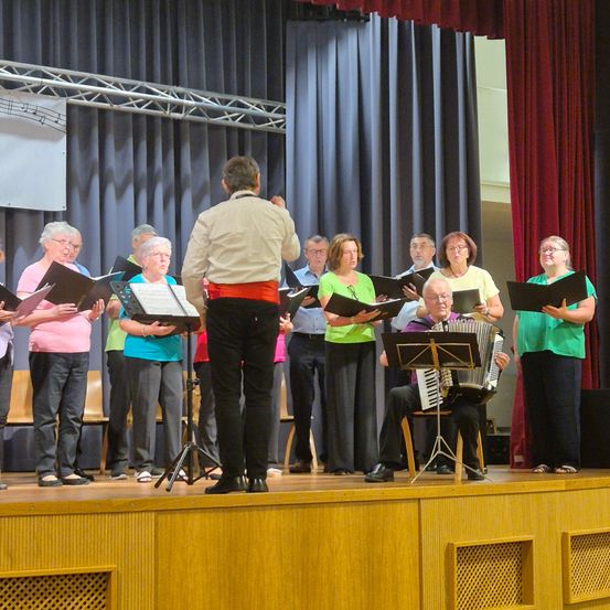 Eine Gruppe älterer Erwachsener auf der Bühne singt mit einem Mann, der ein Akkordeon spielt. Der Mann vorne steht mit einem offenen Buch. Hinter ihnen sind rote Vorhänge.