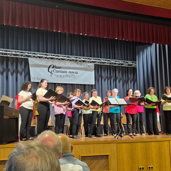 Ein Chor namens 'Chor Cantate nova' tritt auf der Bühne auf. Sie halten Musikbücher in den Händen. Ein Publikum beobachtet sie.