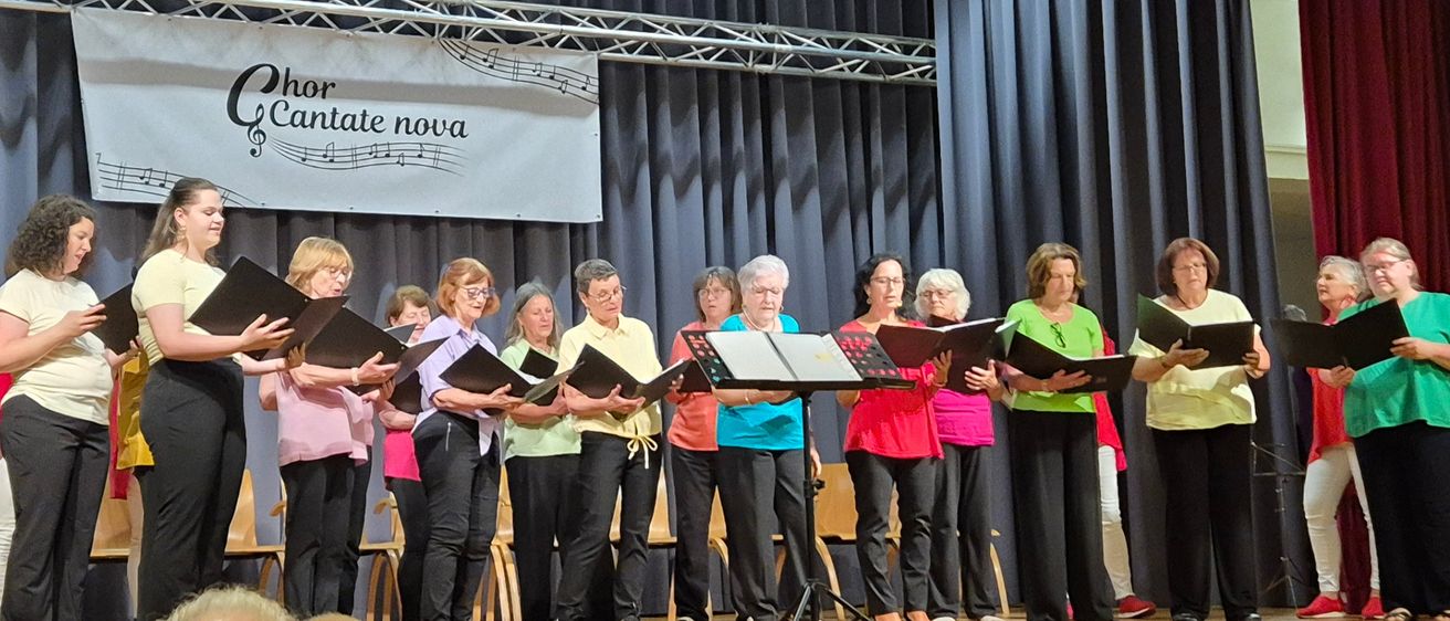 Ein Chor namens Chor Cantate Nova steht auf der Bühne, hält Notenblätter und singt. Ein Banner mit Musiknoten und Text steht dahinter. Ein Klavier befindet sich links. Zuschauer sind vor ihnen.