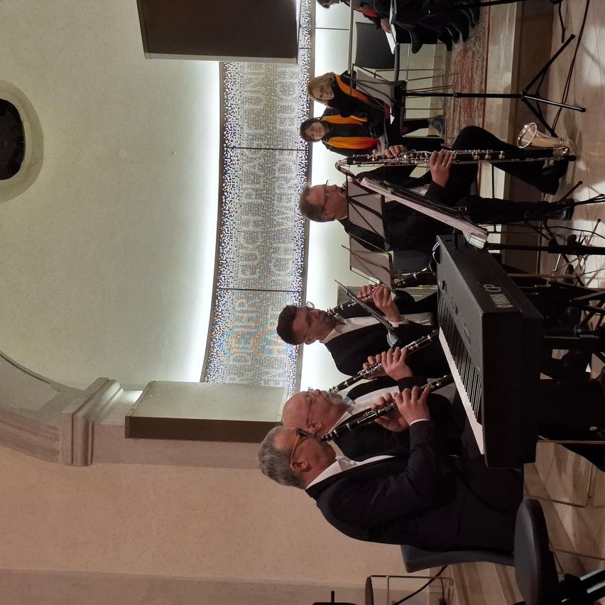 Eine Gruppe von Musikern in Anzügen und mit Brillen spielt Musikinstrumente, darunter Klarinetten und ein Klavier, in einem Raum mit gefliestem Boden und einer Wand mit Design.