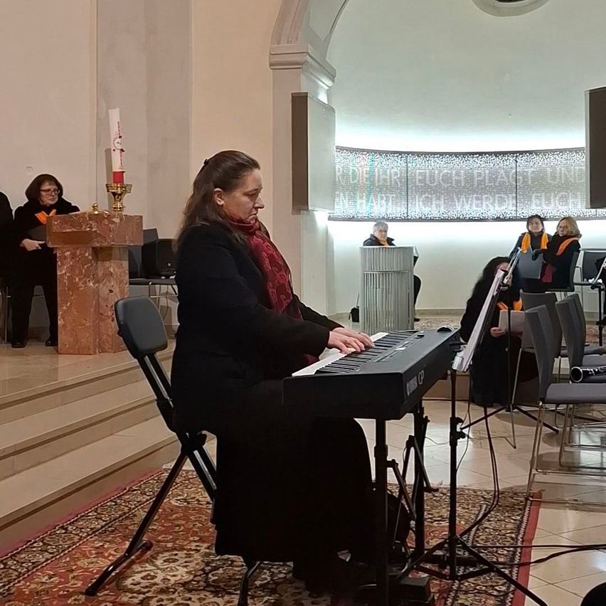 Eine Frau spielt Klavier in einer Kirche, während andere um sie herum sitzen. Der Raum hat einen Marmoraltar und eine Kerze.