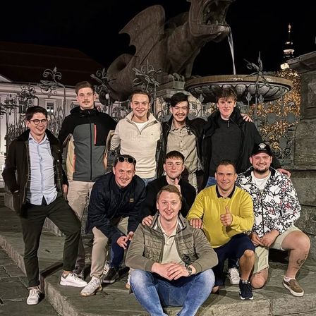 Eine Gruppe von Männern posiert nachts vor einem Brunnen mit Drachenstatue für ein Foto. Sie lächeln und tragen verschiedene Kleidungsstücke.