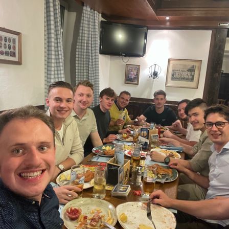 Eine Gruppe von Männern lächelt und posiert für ein Foto an einem Tisch in einem Restaurant. Sie haben verschiedene Speisen auf ihren Tellern, Gläsern und Servietten.