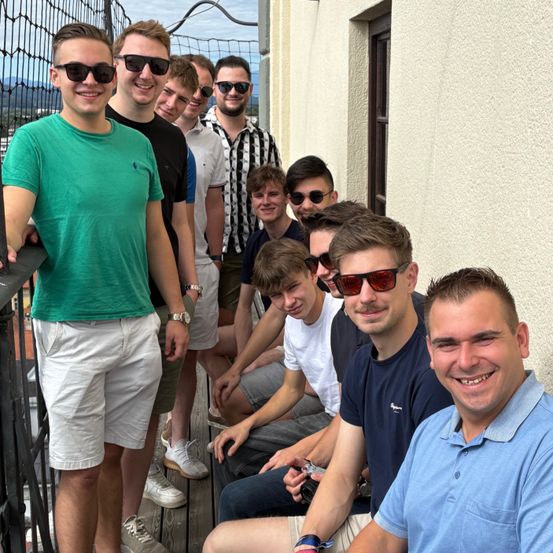 Eine Gruppe von Männern steht auf einem Balkon, einige sitzen auf dem Boden, alle mit Sonnenbrillen. Der Mann im grünen Shirt steht links, während andere Freizeitkleidung tragen. Sie scheinen für ein Foto zu posieren.