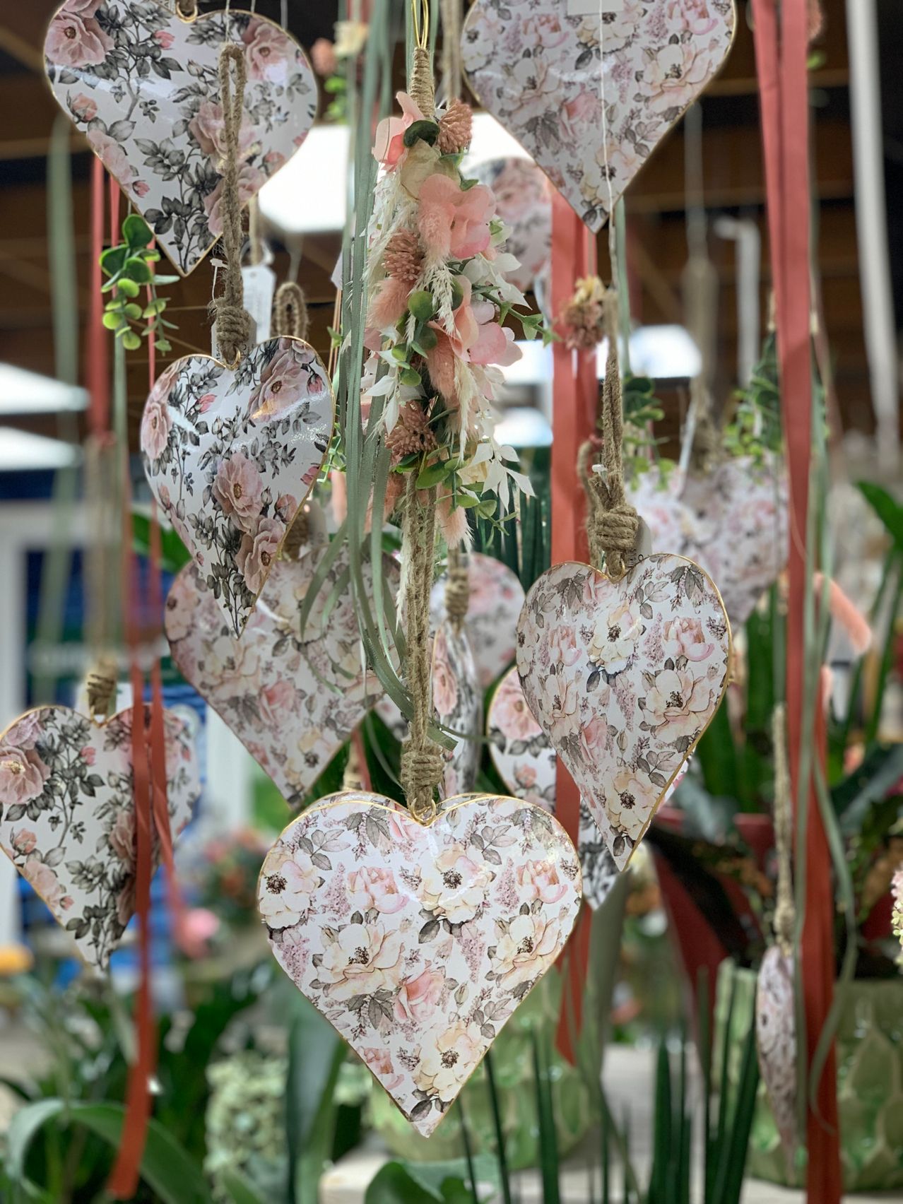 Mehrere herzförmige Dekorationen mit Blumenmustern hängen von der Decke, verziert mit Blumen und grünen Blättern, aufgehängt an roten Bändern in einem gut beleuchteten Raum.