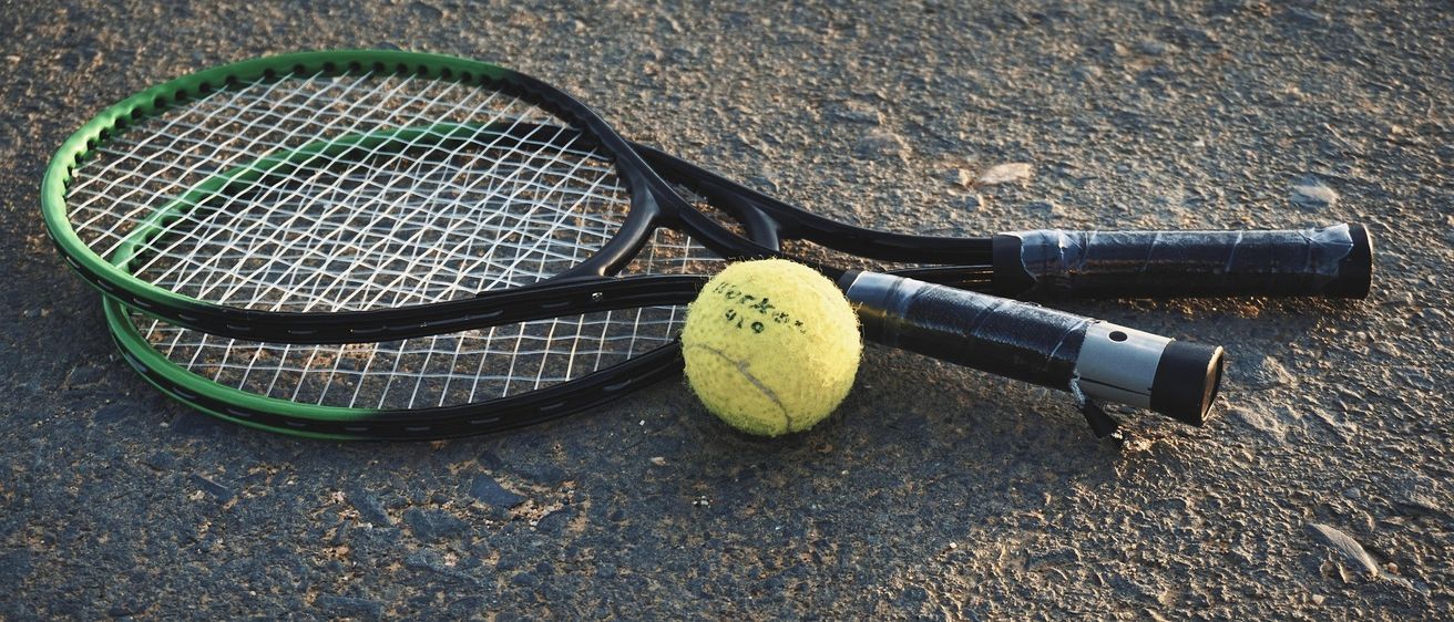 Ein Tennisschläger und ein Tennisball liegen auf einer Betonoberfläche. Der Schläger ist schwarz mit grünen Akzenten und der Ball ist gelb.