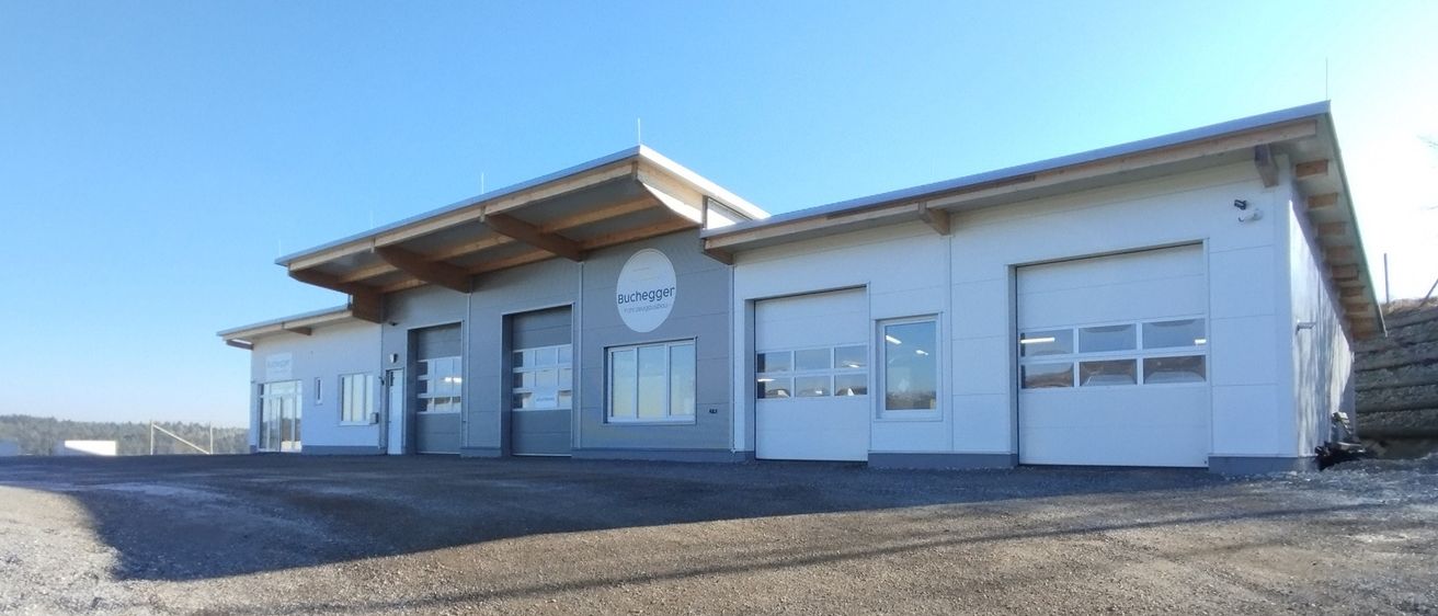Ein modernes Gebäude mit flachem Dach und großen Fenstern befindet sich in einem Kiesparkplatz. Das Gebäude hat mehrere Türen und ein rundes Logo mit dem Text Buchegger.