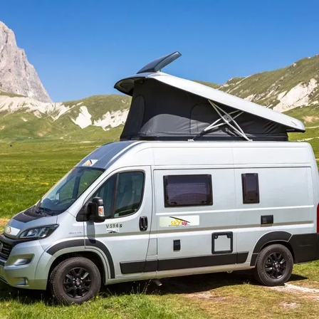 Ein silberner Wohnmobil mit angehobenem Dach steht auf einer Grasfläche mit Bergen im Hintergrund.