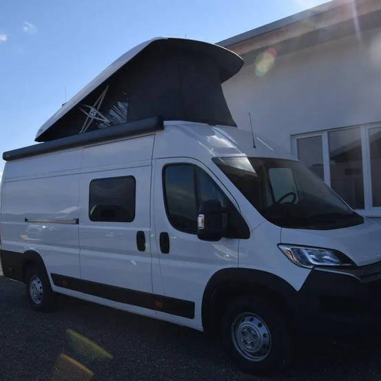 Ein weißer Wohnmobil mit offenem Dach ist vor einem Gebäude geparkt. Der Himmel ist blau mit einigen Wolken.