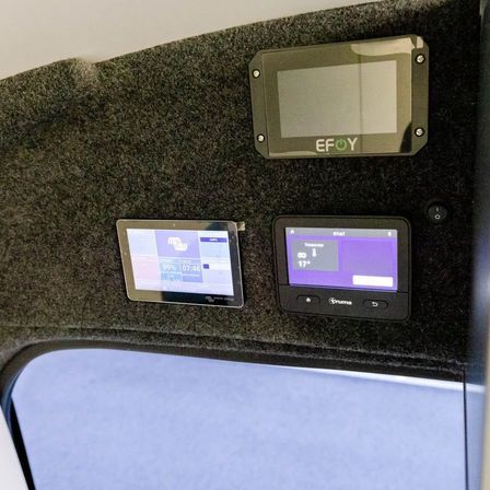 Drei Bildschirme sind an einer schwarzen Oberfläche montiert. Der obere Bildschirm zeigt EFOY. Der mittlere Bildschirm zeigt einen Batterieprozentsatz von 99% und eine Zeit von 07:46. Der untere Bildschirm zeigt 17 Grad Celsius an.