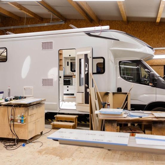 Ein weißer Wohnmobil wird in einer Werkstatt repariert. Die Tür steht offen und zeigt den Innenraum, der einen Küchenbereich enthält. Verschiedene Werkzeuge und Materialien sind herum verstreut.
