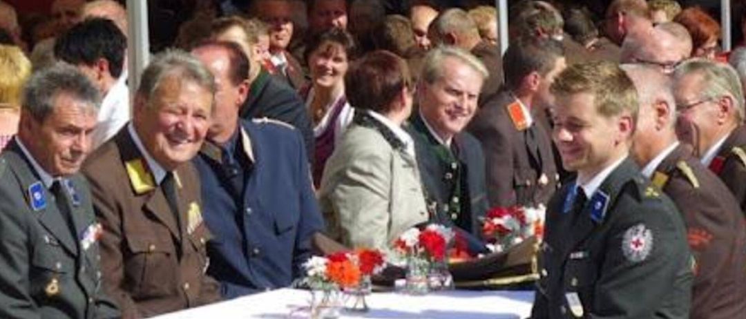 Eine Gruppe von Menschen, einige in Uniformen, versammelt sich um einen Tisch mit Blumen, wahrscheinlich bei einer Zeremonie.