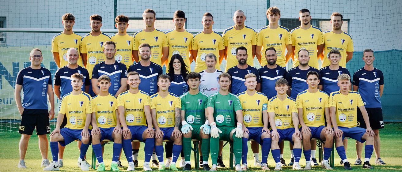 Das Fußballteam posiert für ein Foto auf dem Feld mit einem weißen Banner, das 'für einen' in roten Buchstaben steht. Die Teammitglieder tragen blaue und gelbe Uniformen mit verschiedenen Sponsorenlogos.