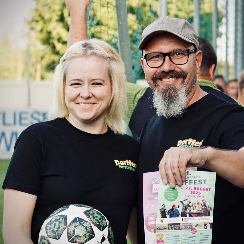 Eine lächelnde Frau und ein Mann bei einem Fußballevent, die einen Fußball und eine Broschüre halten, mit verschwommenen Menschen und einem Zaun im Hintergrund.