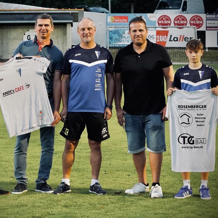 Vier Männer und ein Junge halten Trikots auf einem Rasenfeld. Hinter ihnen sind Banner angebracht, eines mit einem Logo und Text. Der Mann links hält ein Trikot mit den Worten 'ANDI'S CAFE'.