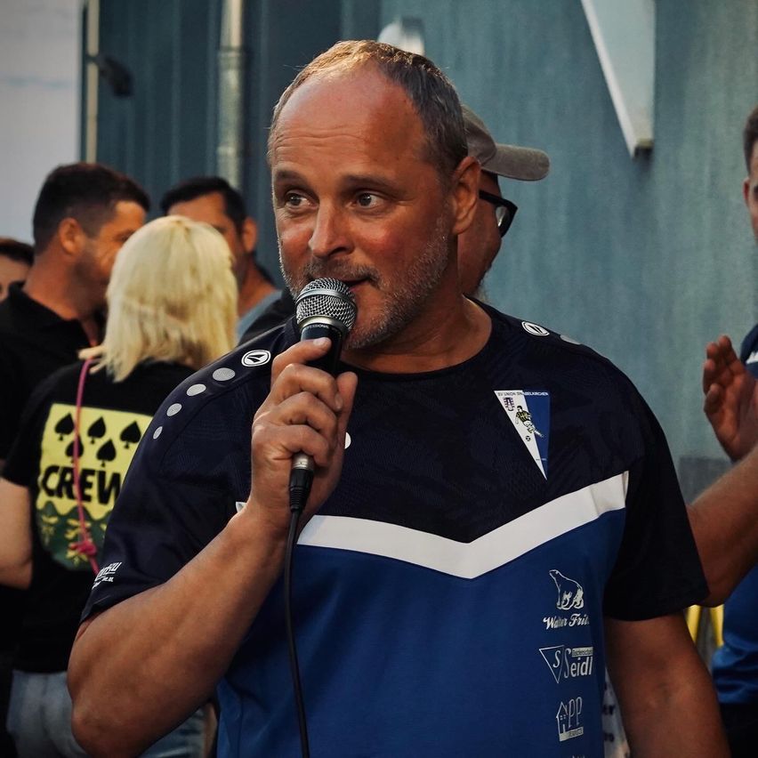 Ein Mann mit einem Mikrofon steht vor einer Menge und trägt ein blaues Shirt mit weißen Akzenten und einem Logo auf der linken Seite.