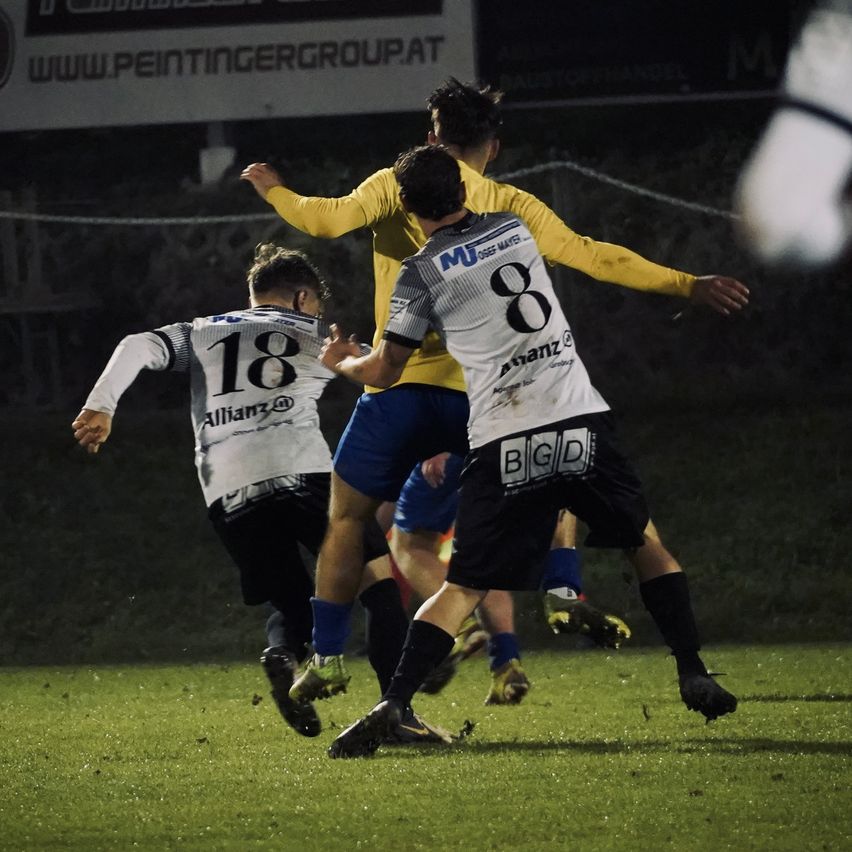 Drei Fußballspieler sind auf einem Feld. Sie tragen Trikots mit den Nummern 8, 18 und einer anderen Nummer. Der Spieler in der Mitte trägt ein gelbes Trikot.