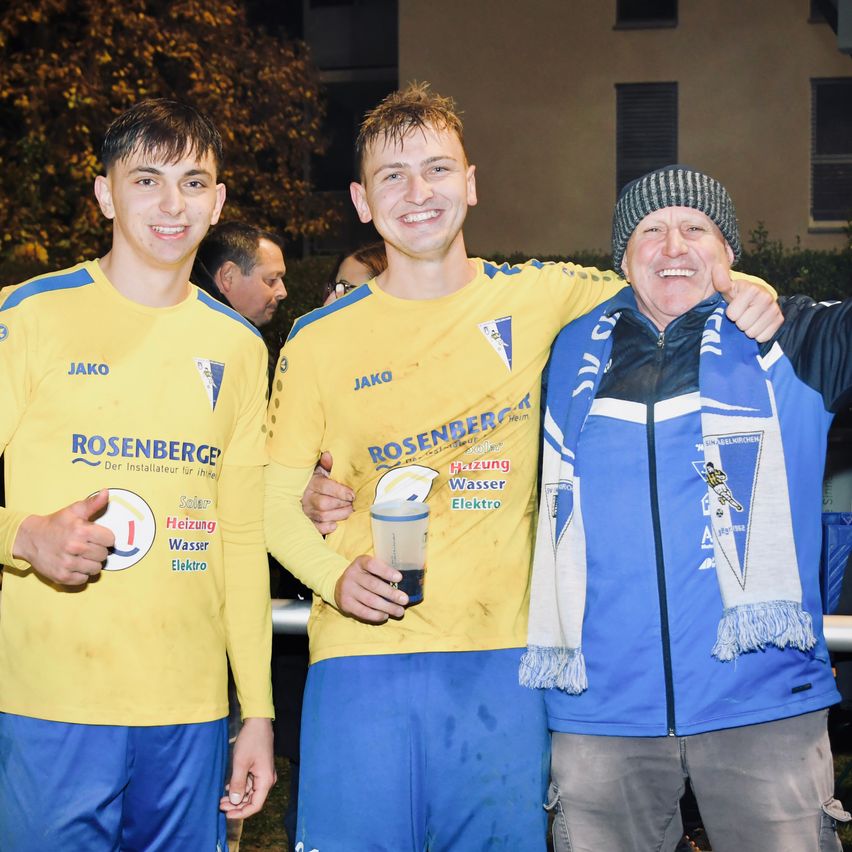 Drei junge Männer in Sporttrikots stehen lächelnd zusammen. Der Mann in der Mitte hält einen Becher und hat seinen Arm um die anderen beiden gelegt. Der Mann auf der rechten Seite trägt eine Mütze und ein Halstuch.