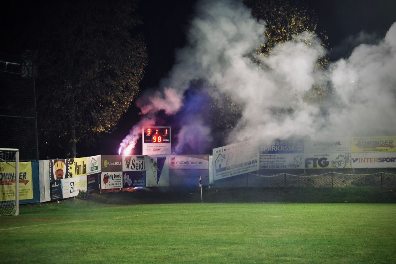 Rauch steigt auf einem Sportplatz auf, ein Anzeigetafel zeigt 98 verbleibende Minuten. Plakatwände säumen den Rand, und Bäume sind im Hintergrund sichtbar.