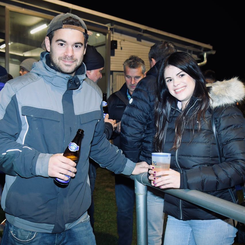 Eine Gruppe von Menschen steht an einem Geländer, ein Mann hält eine Flasche und eine Frau einen Becher. Sie sind draußen bei Nacht, der Mann und die Frau lächeln.