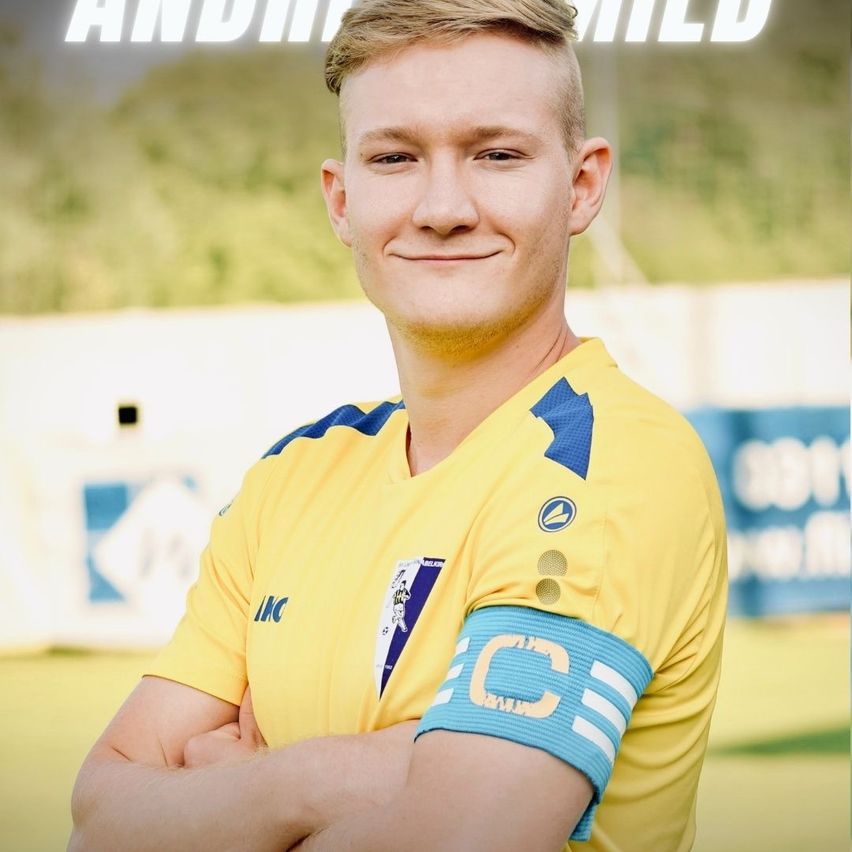 Ein lächelnder Fußballspieler in einem gelben Trikot mit blauen Akzenten und einem blauen Armband mit dem Buchstaben 'C' steht auf einem Feld mit unscharfem Hintergrund.
