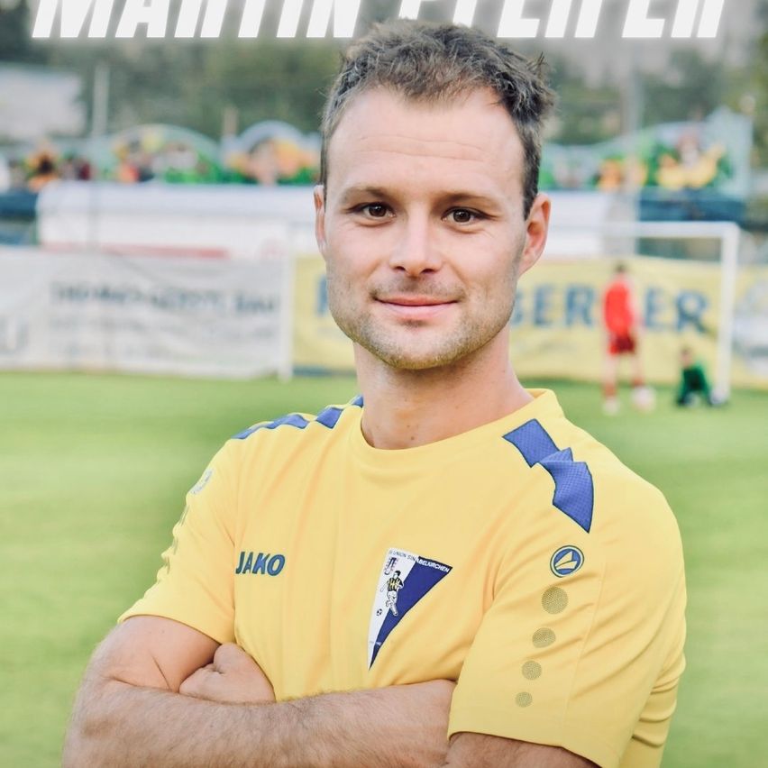Ein Mann in einem gelben Fußballtrikot mit 'Jako' bedruckt, lächelt und steht mit verschränkten Armen auf einem Fußballfeld.