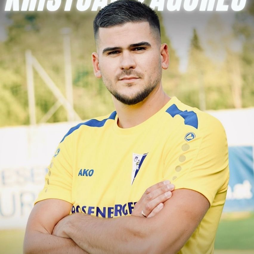 Ein Fußballspieler in einem gelben Trikot mit blauen Akzenten posiert mit verschränkten Armen auf einem Feld. Der Hintergrund ist unscharf mit Bäumen und einem Banner.