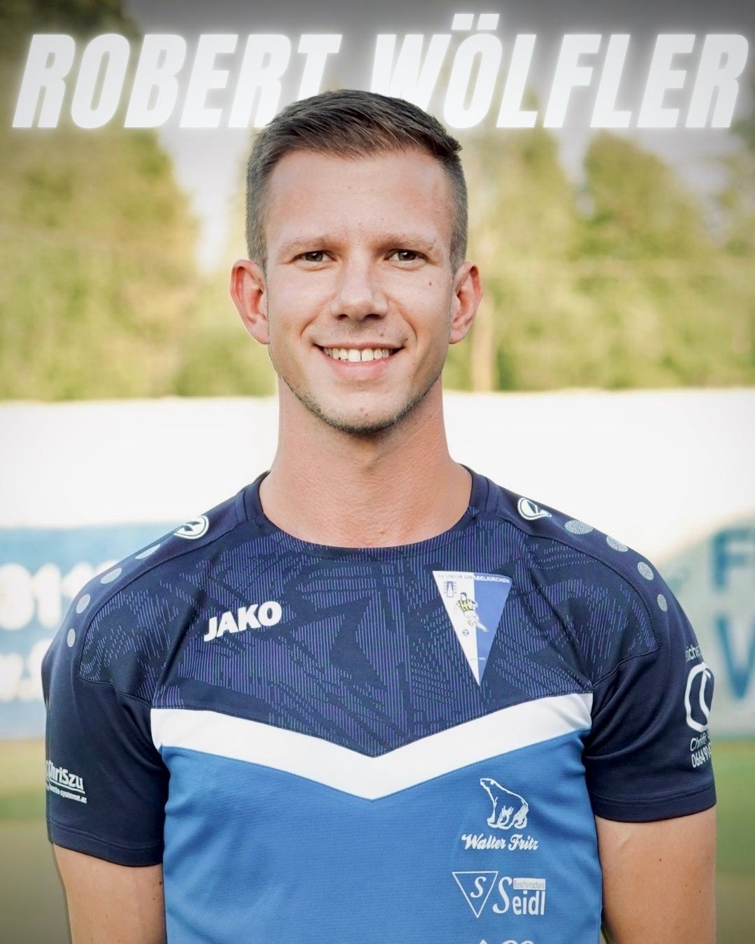 Ein lächelnder Mann in einem blau-weißen Sporttrikot mit 'Jako' auf der Vorderseite und einem Logo auf der linken Brust. Er steht auf einem Feld.